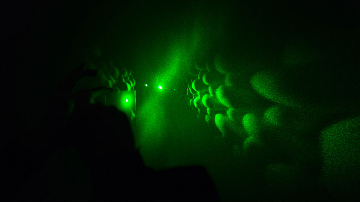green laser light projected on a wall through a glass. The projection on the wall depicts a three dimensional image of woven structure
